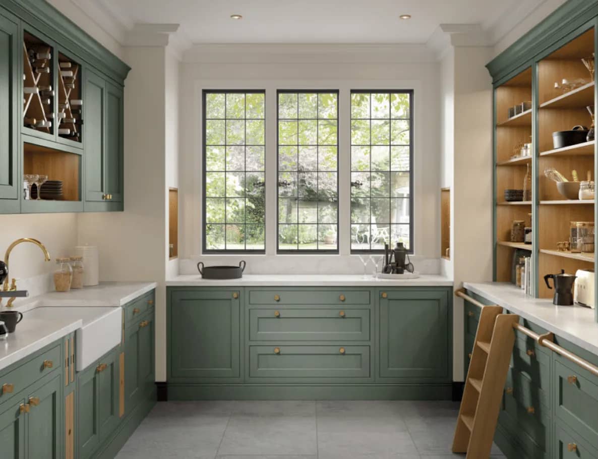 Sage green pantry with a butler sink, brass tap, open oak shelving and a garden window.