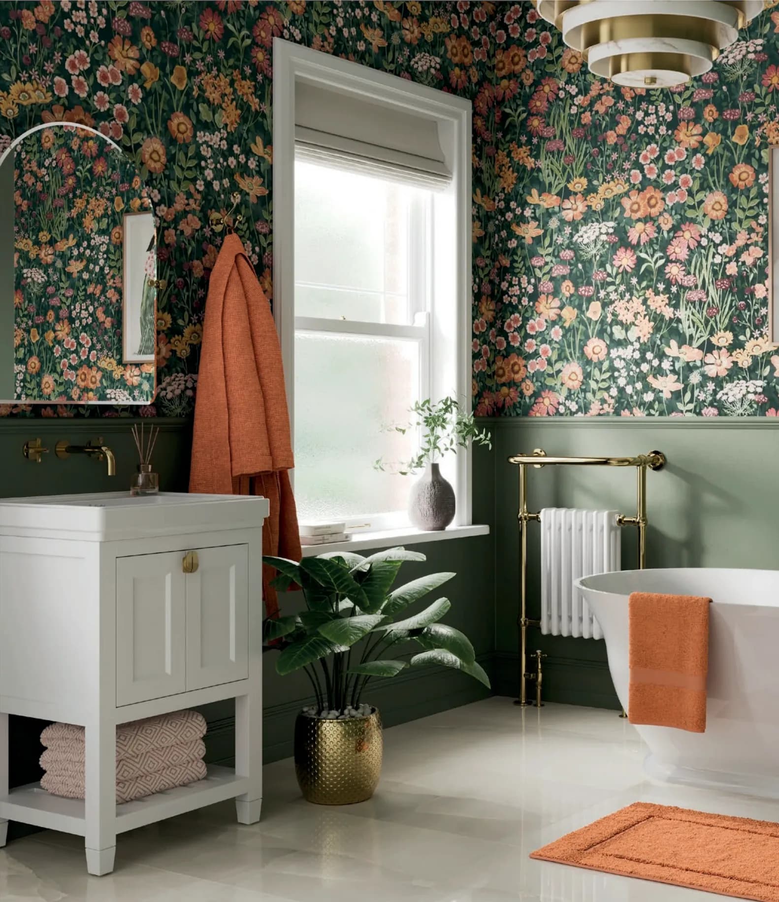 A freestanding bath beside a brass column towel rail, sage green panelling below a floral papered wall, and a white vanity with a wall-mounted brass tap