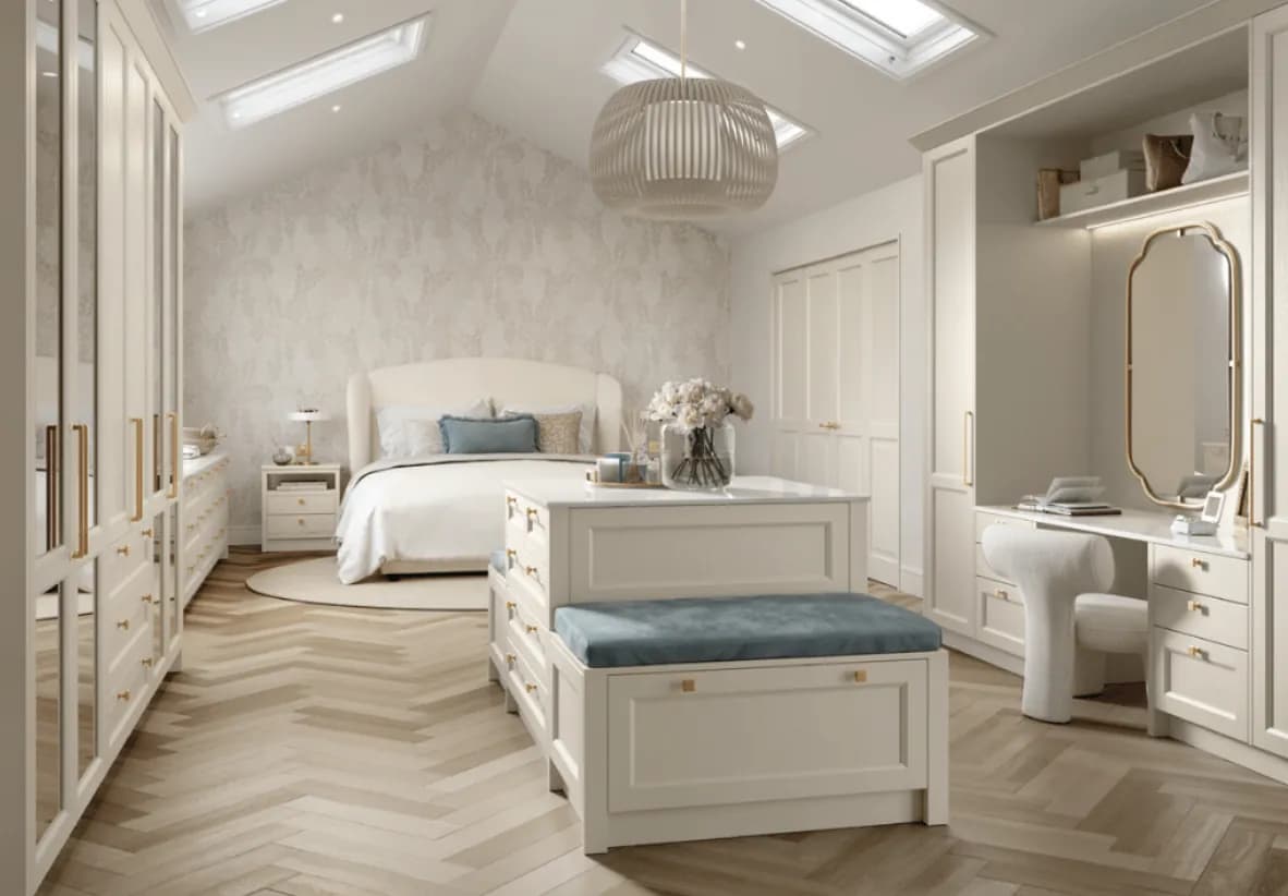 A cream dressing room under roof lights: mirrored wardrobe doors down one side, a dressing table with a lit arched mirror, and an island of drawers with an upholstered box